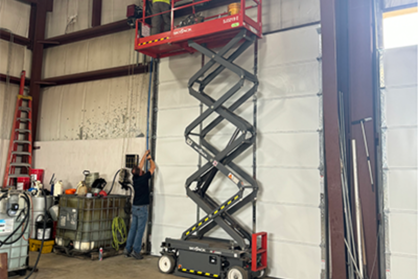 Scissor lift raised beside a warehouse wall, with a worker standing below in an industrial bay.