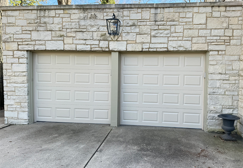 Two beige garage doors in a stone exterior wall with a lantern above and a concrete driveway