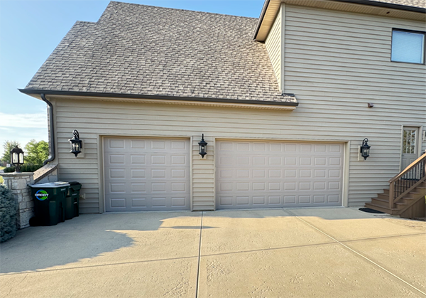 Suburban house exterior with two white garage doors, tan siding, and a concrete driveway