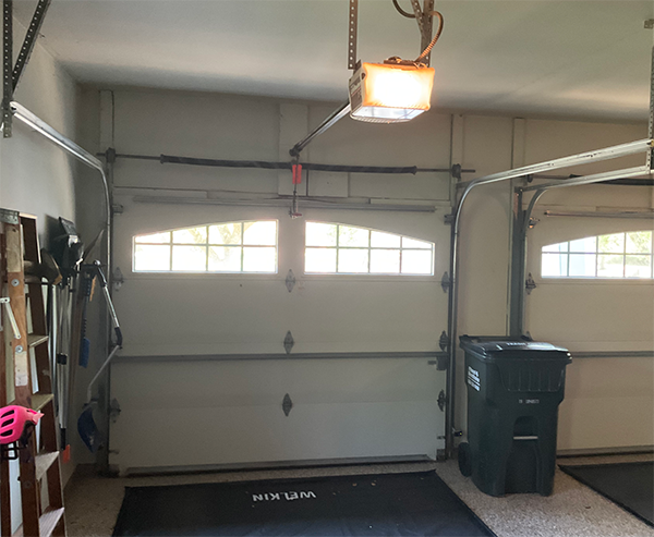 Garage interior with closed white door, overhead opener, and trash bin on the right.