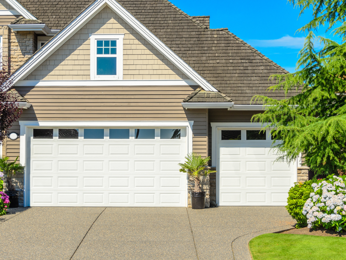 Suburban house exterior with beige siding, white garage doors, and flowering shrubs by the driveway