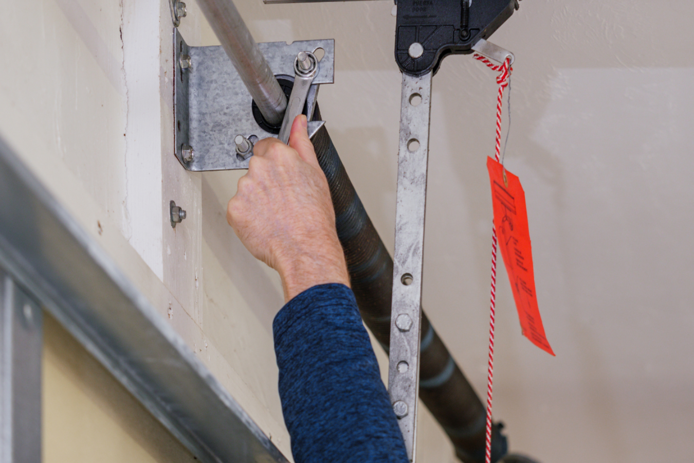 Hand using a ratchet on a wall-mounted bracket with a red safety tag hanging nearby