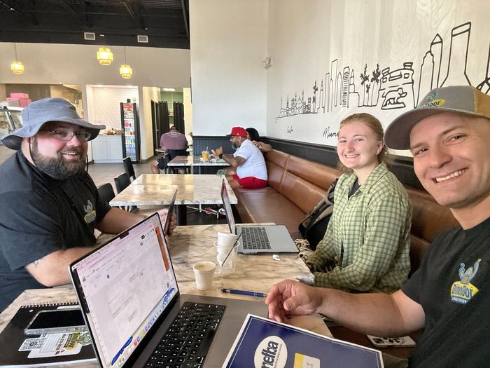 Three people seated at a café table with laptops, smiling and working in a bright restaurant.