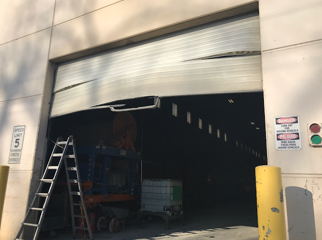 Loading dock with partially open garage door, ladder, pallet jack, and safety signs at a warehouse entrance