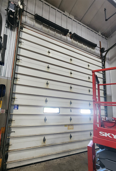 Closed white industrial garage door in a warehouse, with a red lift partially visible on the right.