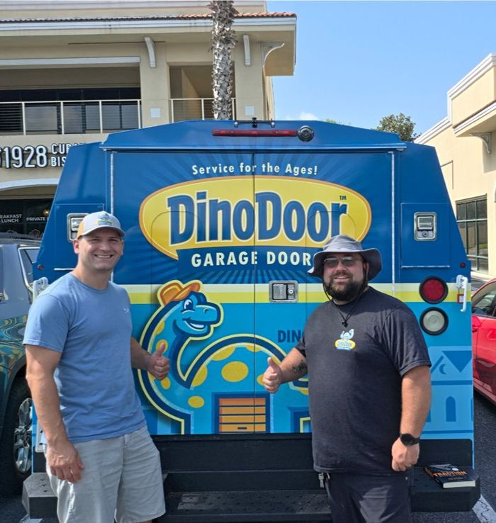 Two men standing in front of a blue DinoDoor garage door service van with dinosaur graphics.