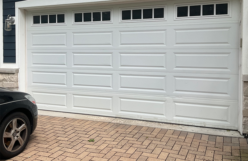 White double garage door with small windows, next to a black car on a brick driveway.