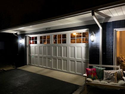 White garage door lit at night beside a lit porch with a cushioned bench.