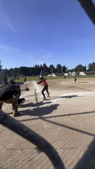 A person wearing a hat and orange shirt, holding a baseball bat on their shoulder on a baseball field.