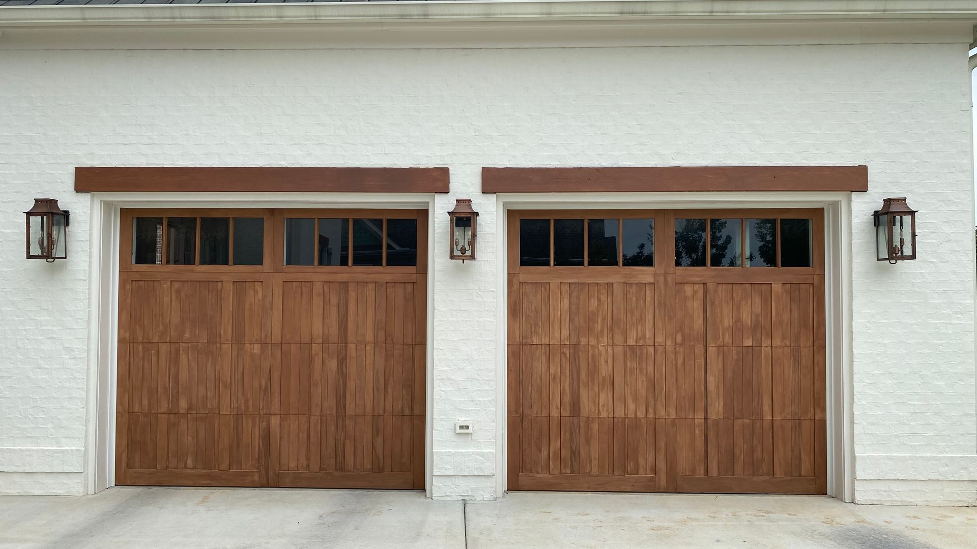 Two wooden garage doors with glass windows, flanked by lanterns on a white brick building.