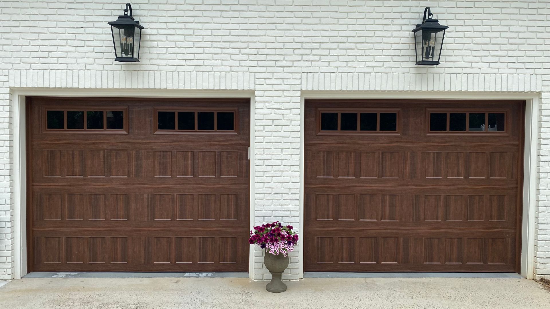 Two brown garage doors on a white brick building with decorative lanterns and a flower pot.