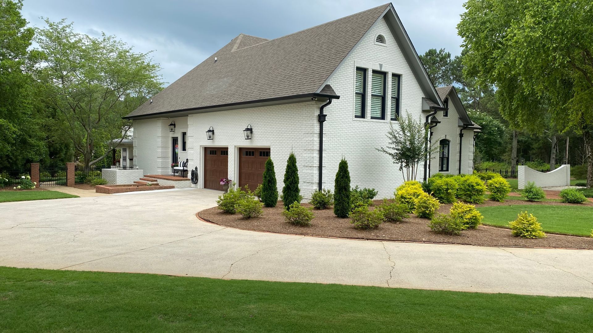 White brick house with brown garage doors and driveway, surrounded by greenery.