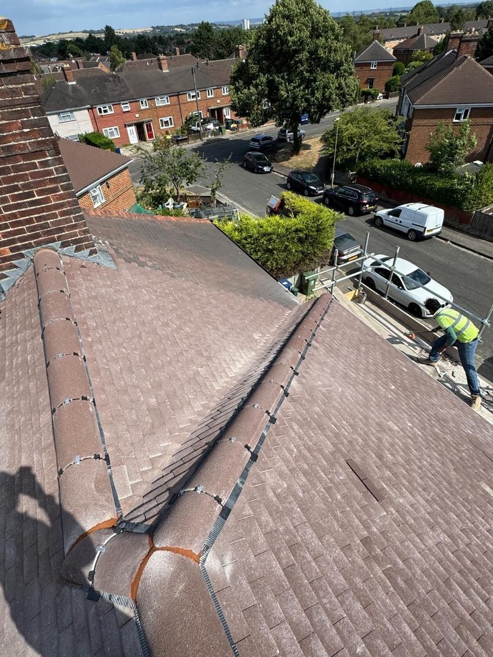A high-angle view of a tiled residential roof with a worker in a high-visibility vest repairing a ridge along the edge.