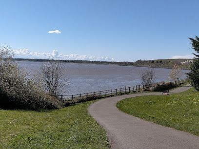 A paved walking path winds through a grassy park toward a calm, wide river under a clear blue sky.