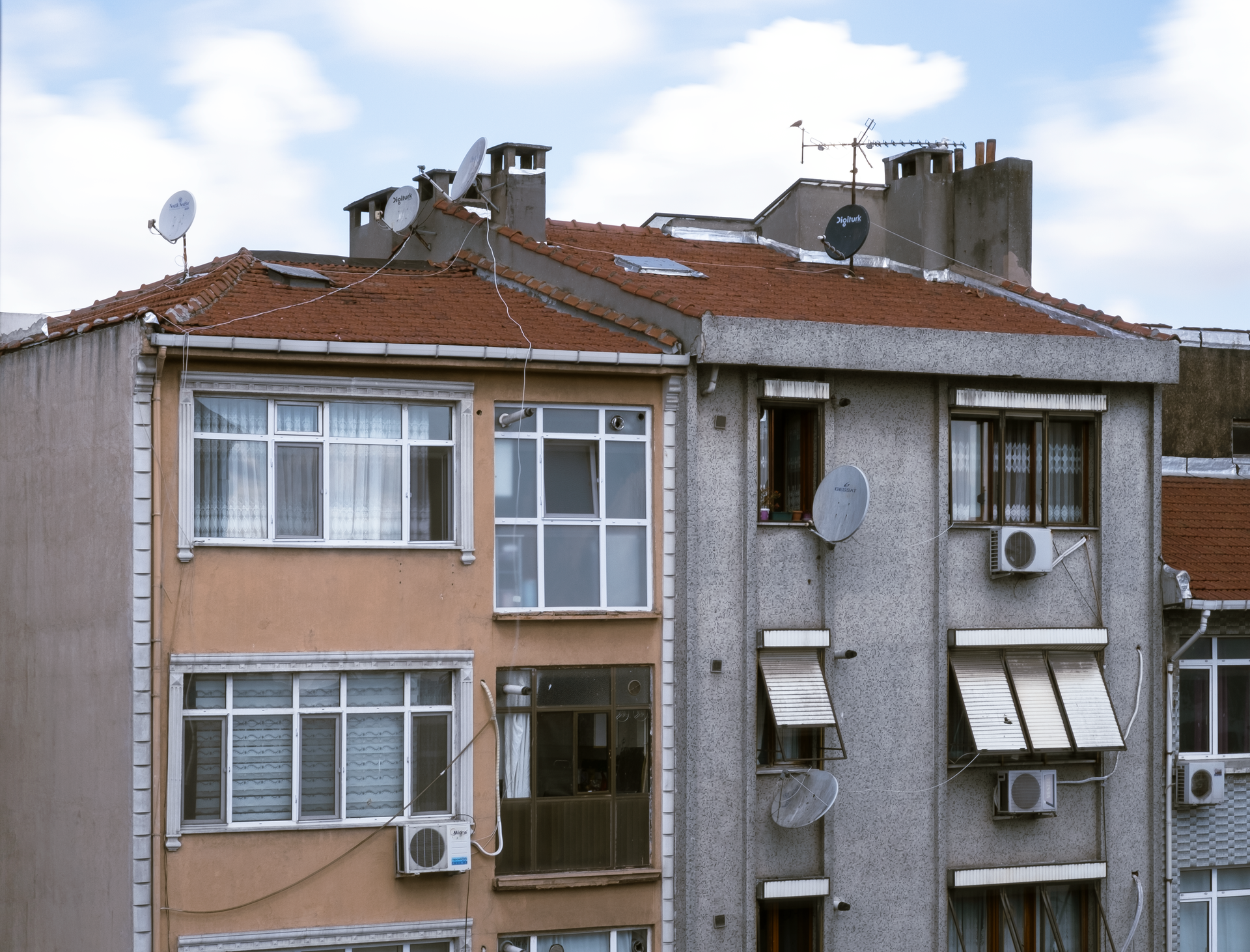 Old apartment building with weathered facades, red-tiled roof, and satellite dishes under a cloudy sky