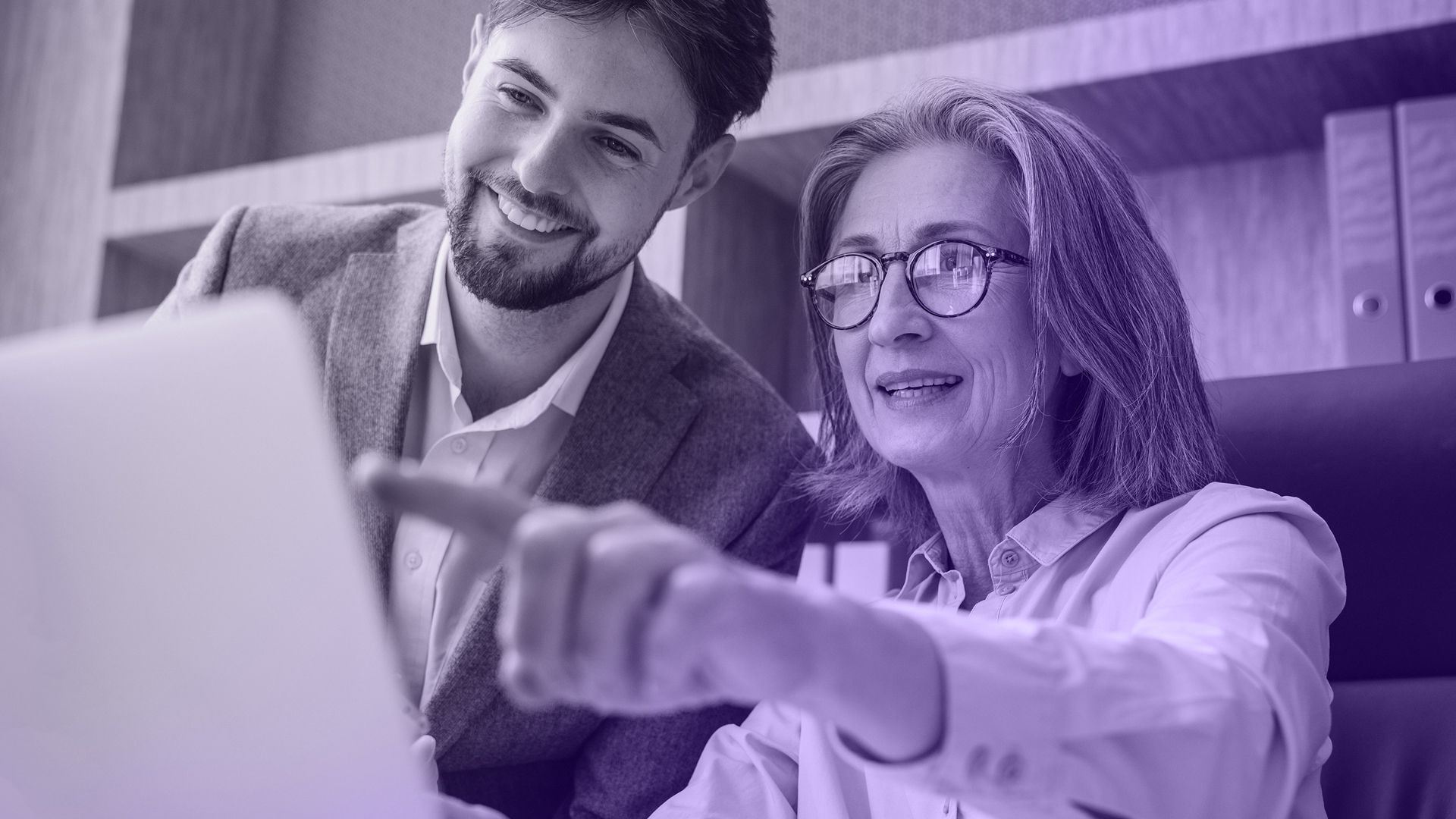 Woman and man looking at a laptop, smiling. The woman points at the screen. Office setting, purple tint.
