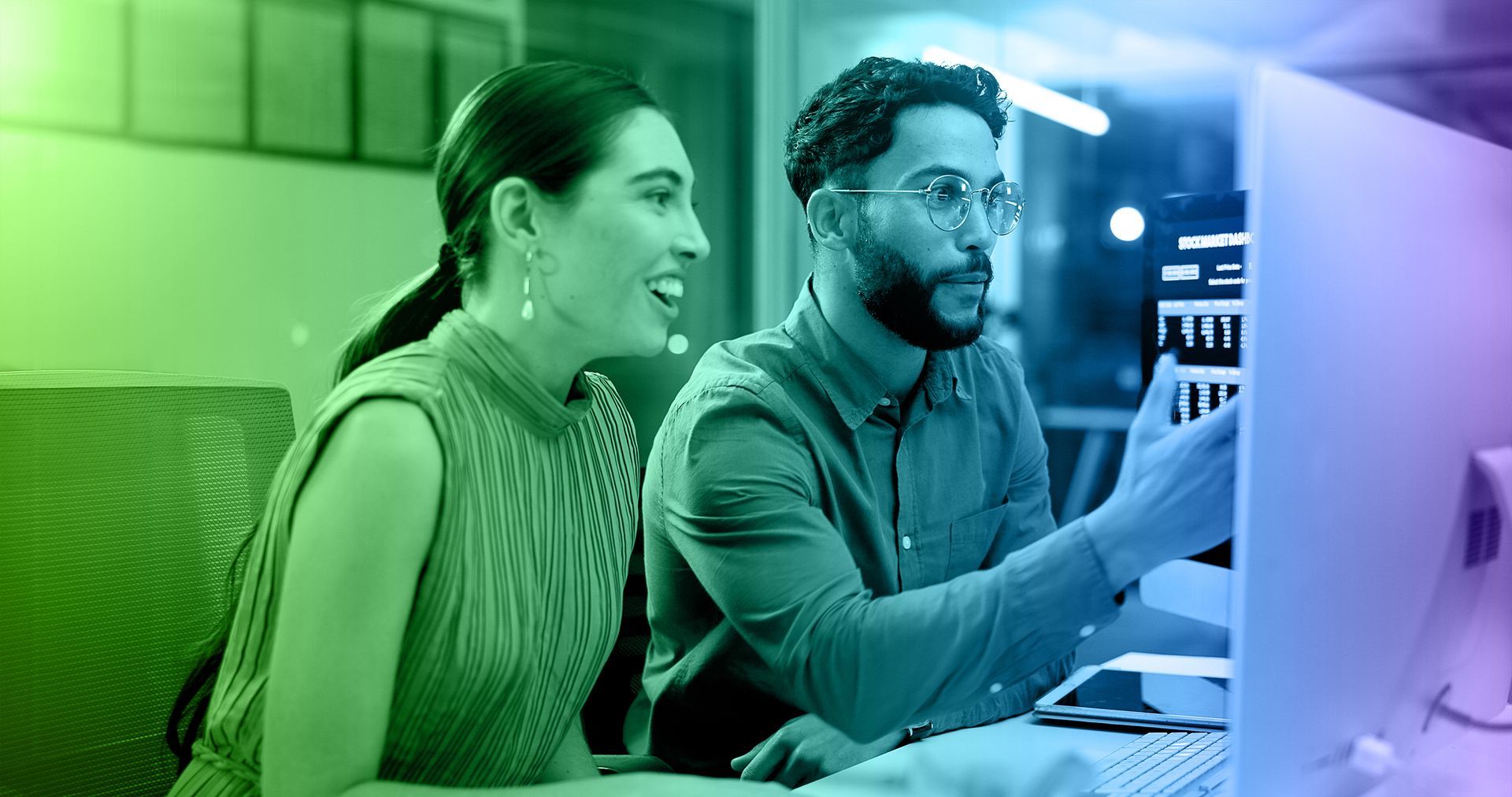 Woman and man looking at a computer screen, discussing the data. Neon color-toned office setting.