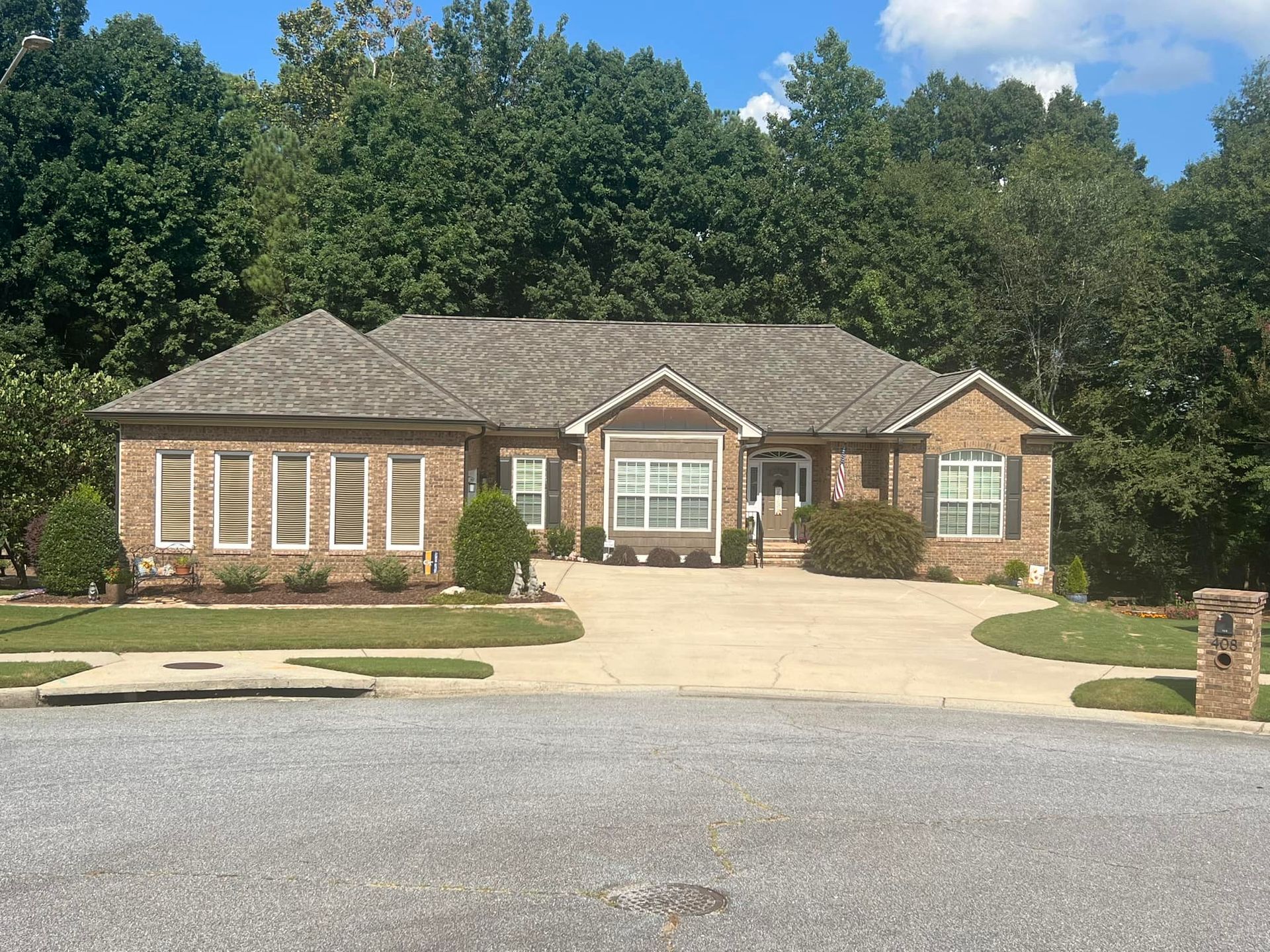 Brick ranch house with brown roof and green lawn, in front of a tree-lined background.