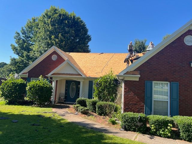 Roofers working on a residential home with exposed wooden roof decking, red brick exterior, and green bushes.