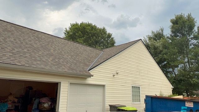 A house with a light-colored exterior and a brown shingled roof, trees, and a cloudy sky.