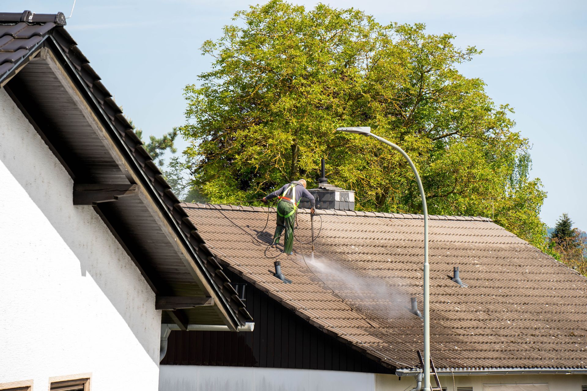 Man in green overalls pressure-washing a brown tile roof; a street lamp and tree are in the background.