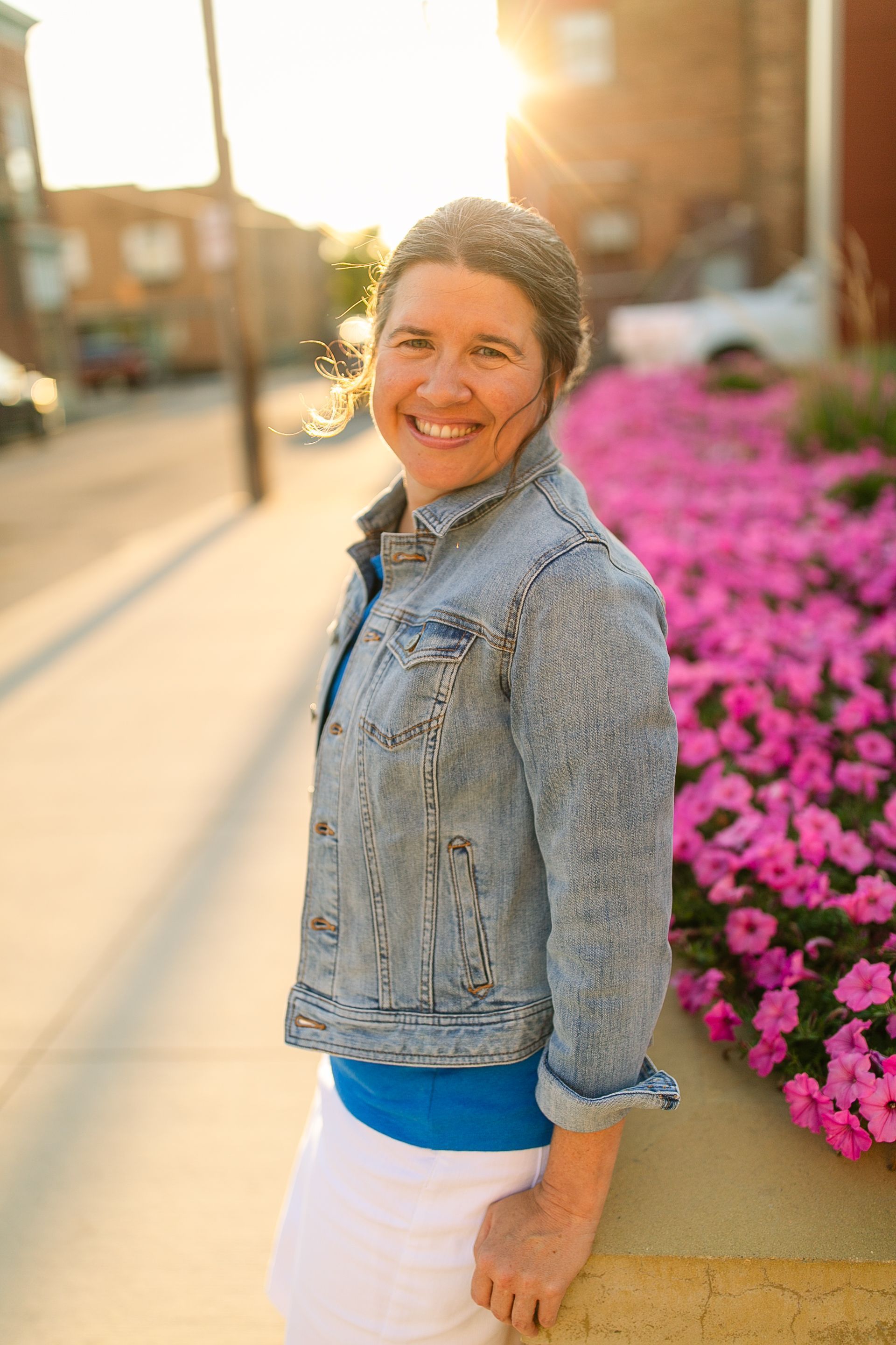 Smiling person in a denim jacket standing beside bright pink flowers on a sunny sidewalk