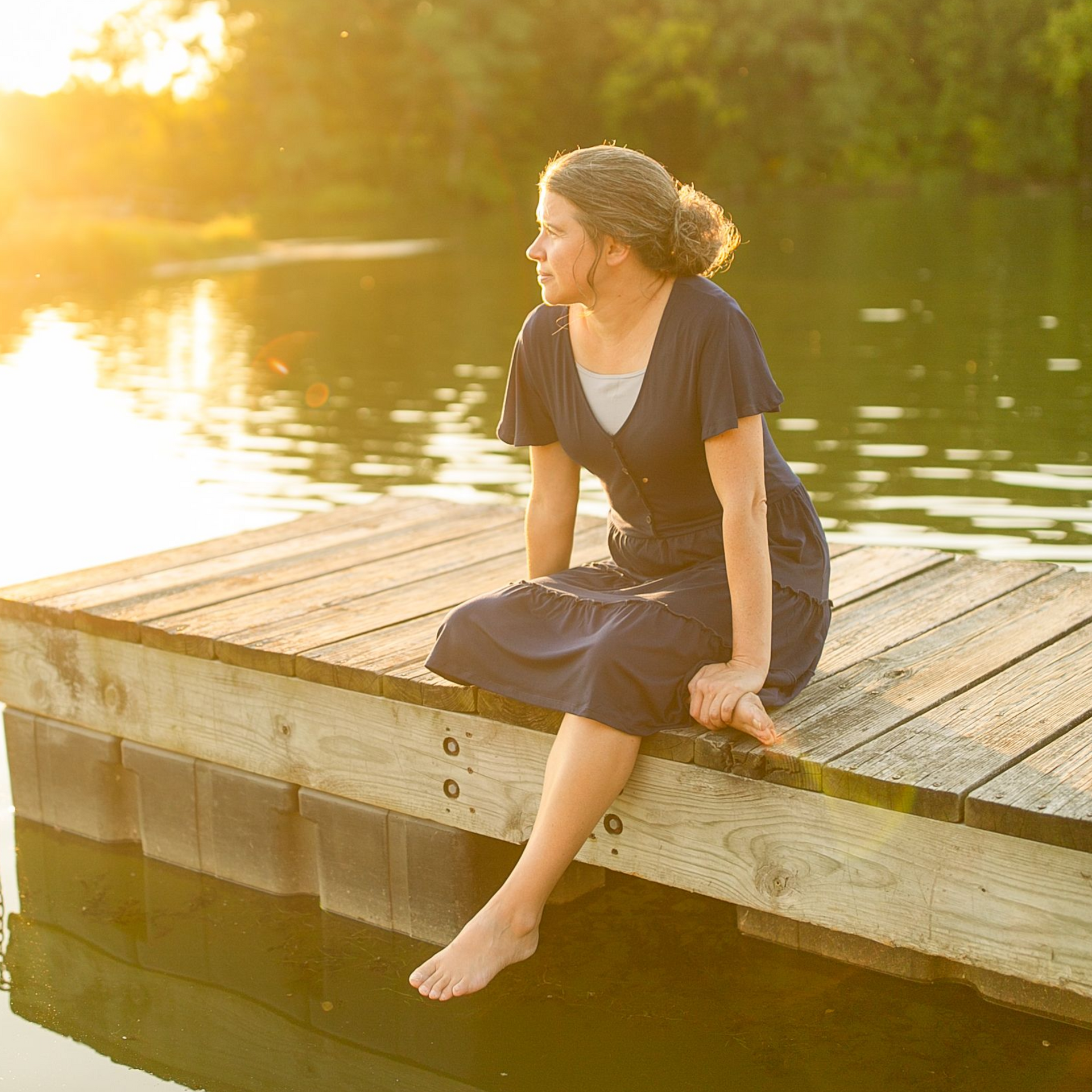 Woman sitting on a wooden dock with bare feet by a calm lake at sunset