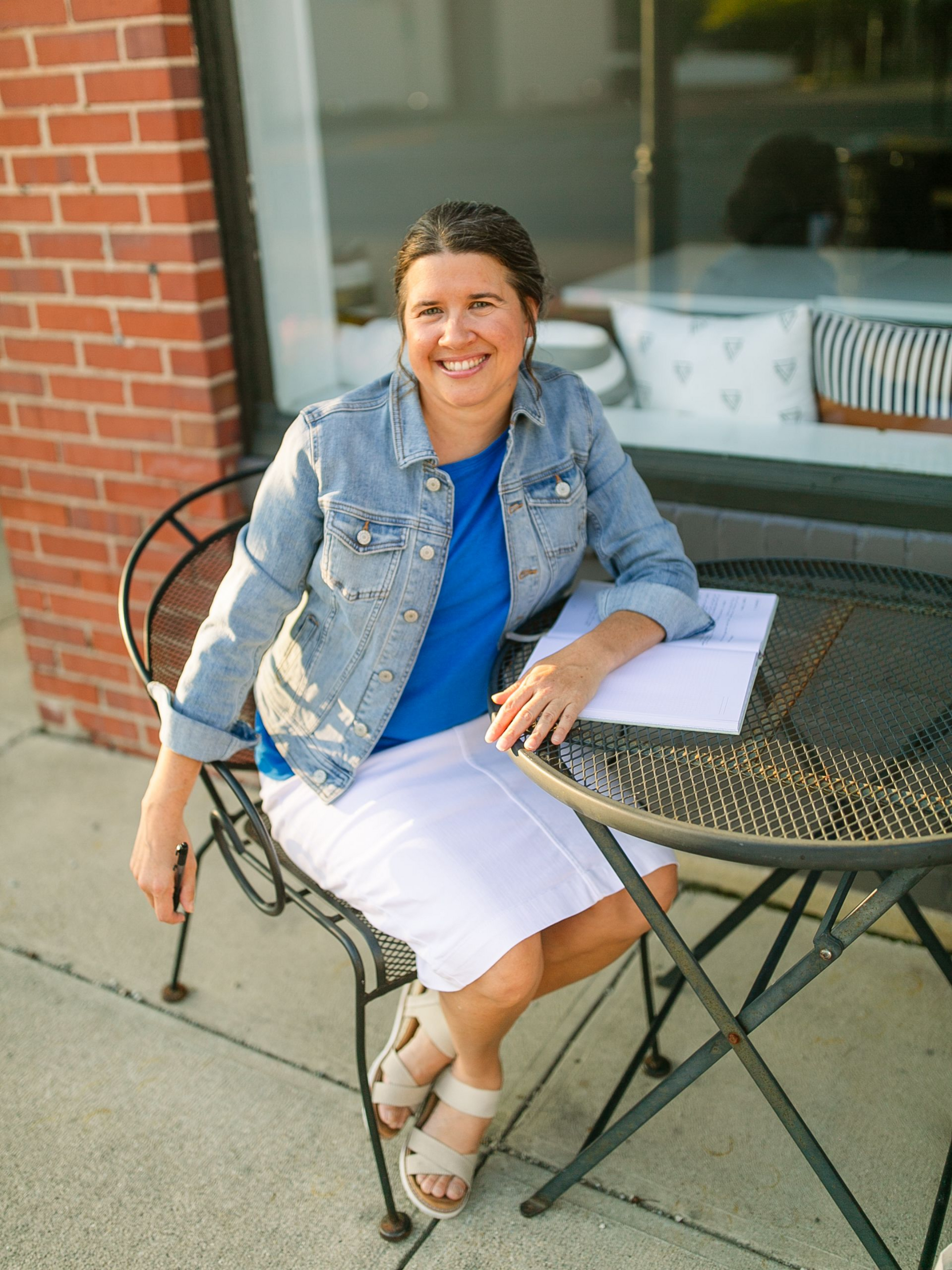 Woman seated at a patio table, smiling, wearing a denim jacket and white skirt beside a brick wall.