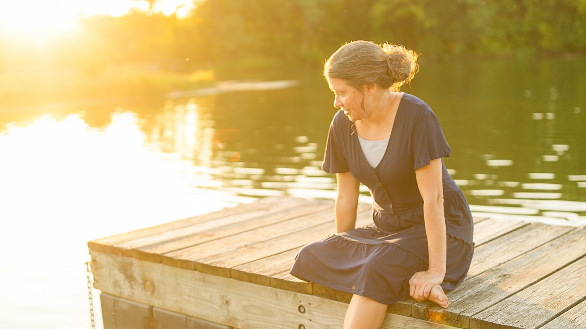Person sitting on a wooden dock by a lake at sunset, with warm golden light reflecting on the water.