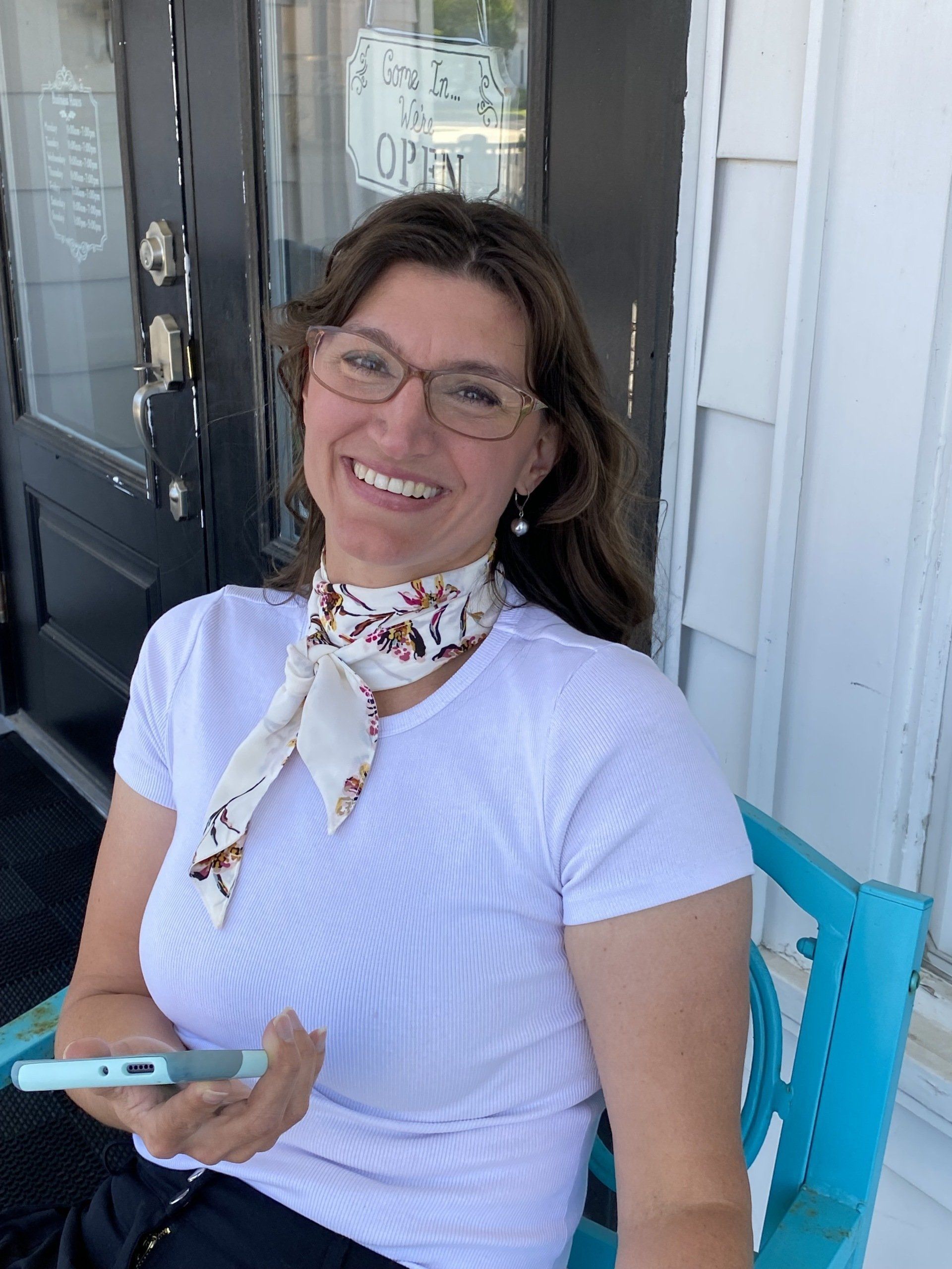 Smiling woman in glasses sitting on a porch, holding a phone, wearing a white shirt and patterned scarf.