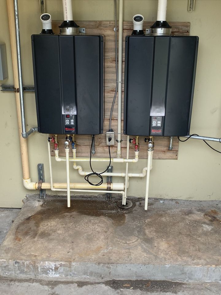 Two black tankless water heaters mounted side by side on an exterior wall with exposed pipes.