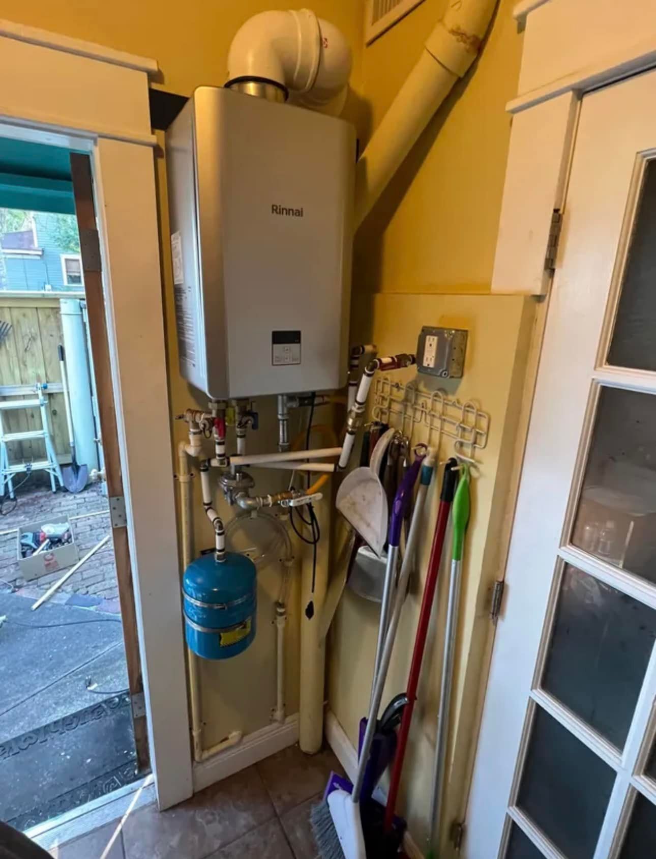 Boiler and utility pipes in a narrow hallway, with brooms and a blue tank mounted on the wall