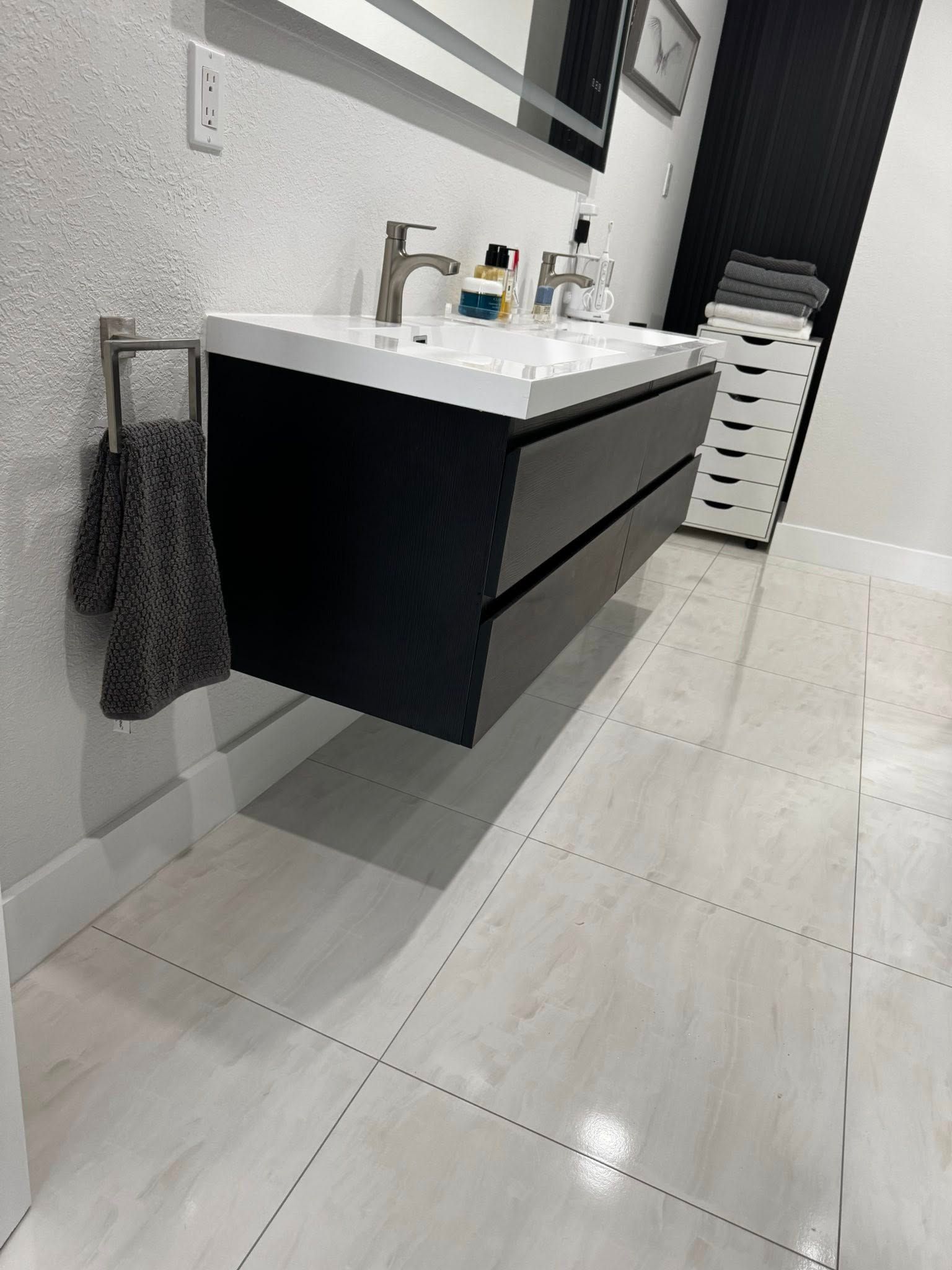 Modern bathroom with floating black vanity, white sink, mirror, and tiled floor; towel hangs on the wall.
