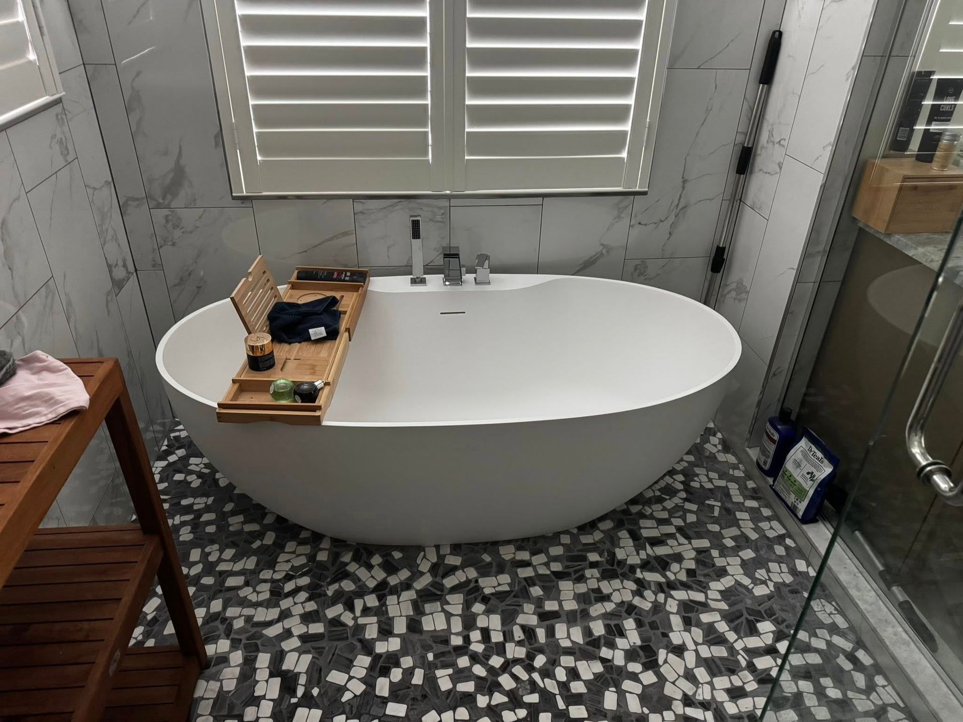 Modern bathroom with a white oval tub, mosaic tile floor, shuttered window, and small bath tray
