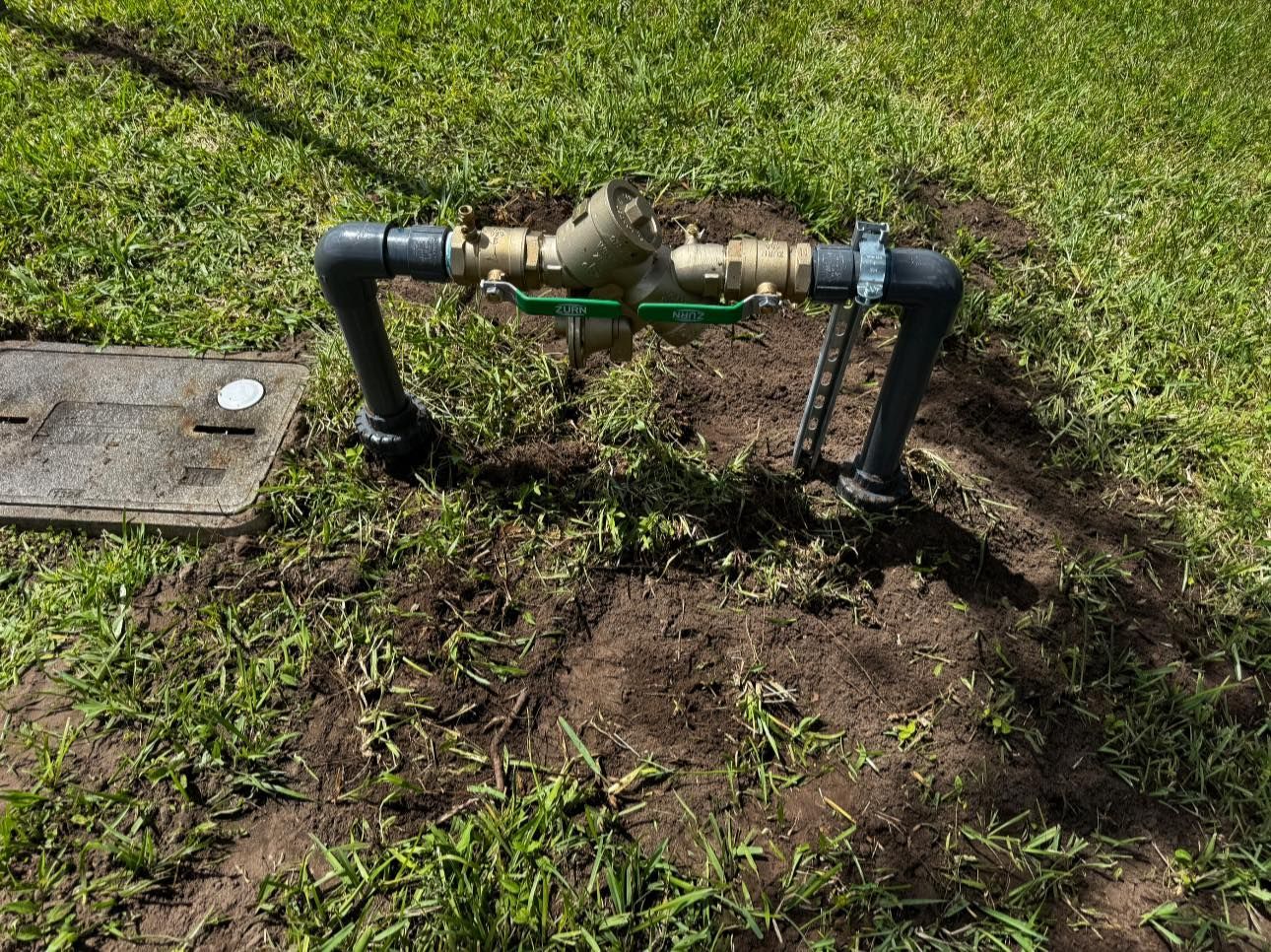 Outdoor water pipe assembly over muddy grass with black PVC pipes and brass valves next to a utility cover
