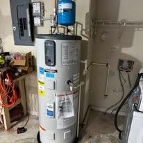 Water heater in a garage with exposed pipes, a blue tank on top, and a cluttered work area nearby