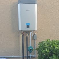 Wall-mounted white water heater with pipes and meter against a beige exterior wall.