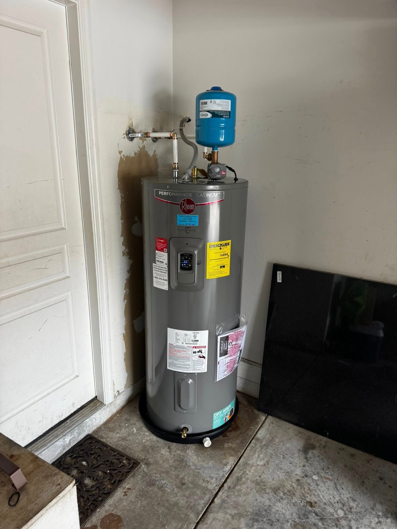 Gray water heater with blue expansion tank in a basement utility corner, beside a white door and black panel