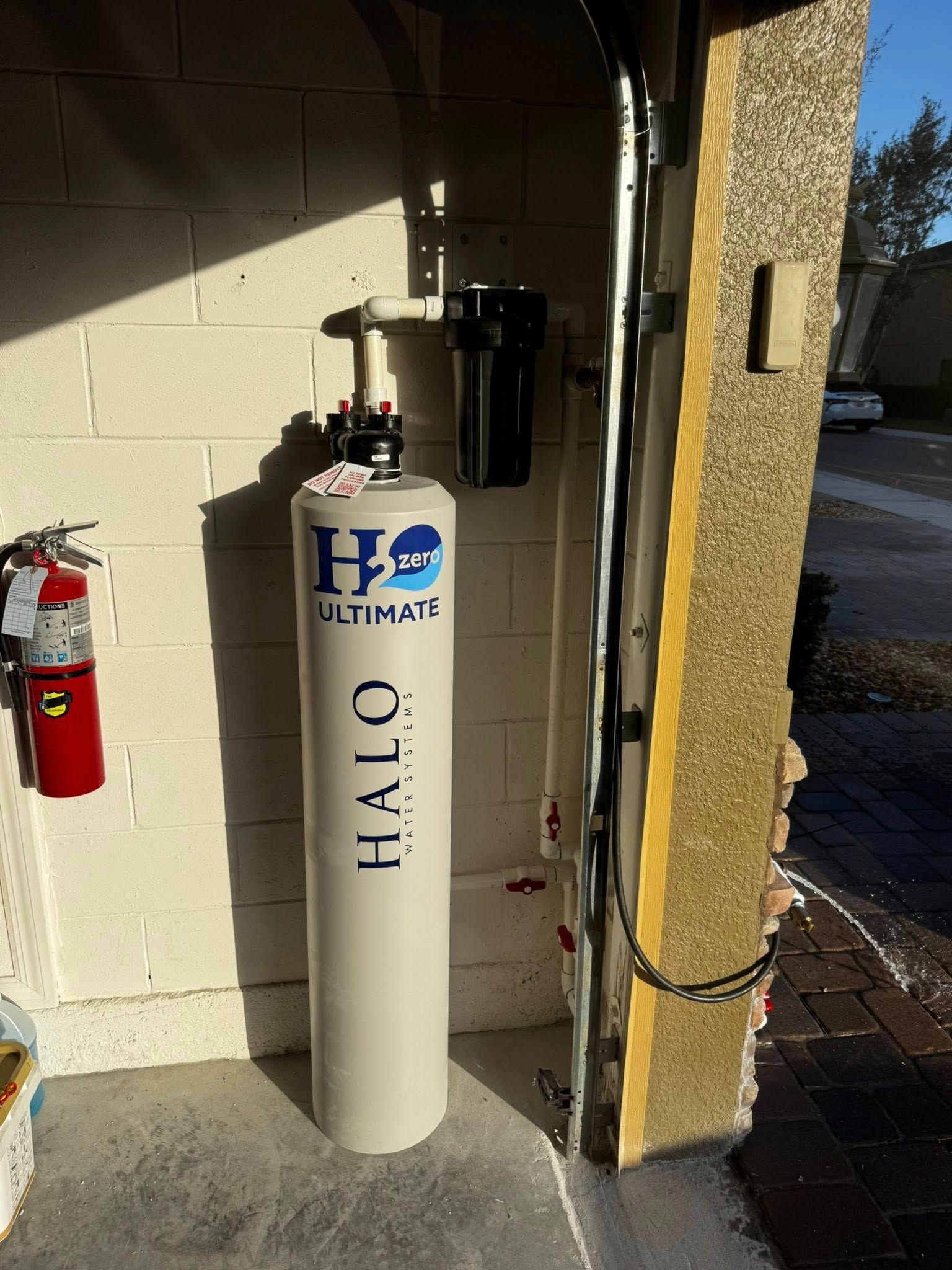 Tall white HEI tank with valves beside a red fire extinguisher in a sunlit exterior alcove