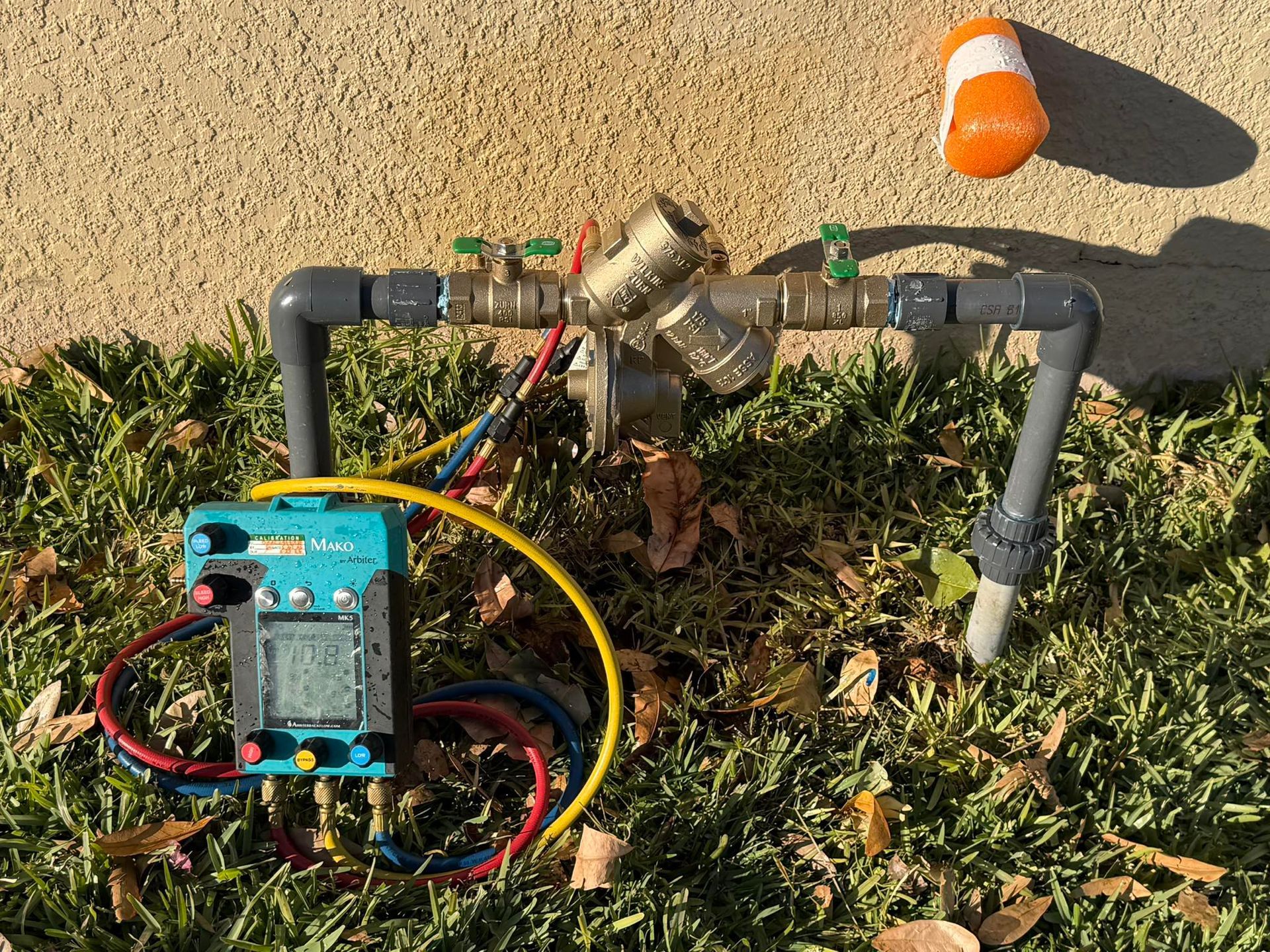 Outdoor utility setup with pipes, valves, wires, and a blue device on grass near a sidewalk