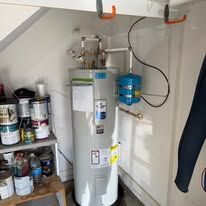 Water heater in a small utility closet with shelves of paint cans and a blue expansion tank attached.