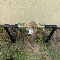 Backflow preventer assembly with black pipes and green valves beside a house wall