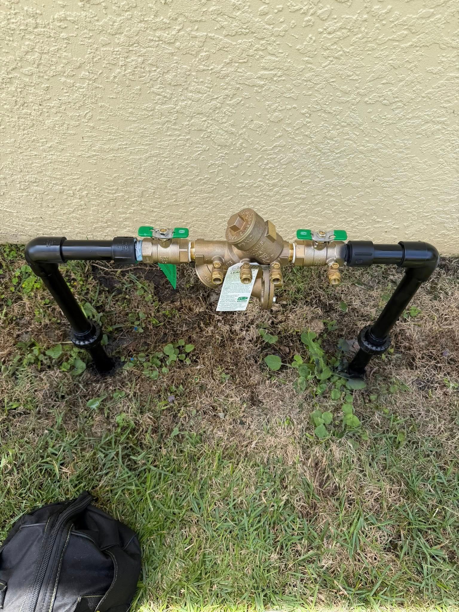 Black metal pipe assembly with two green valves mounted on a wall above grass and dirt