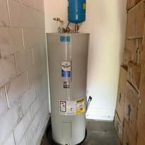 Tall gray water heater in a narrow utility room with white walls and a blue expansion tank on top
