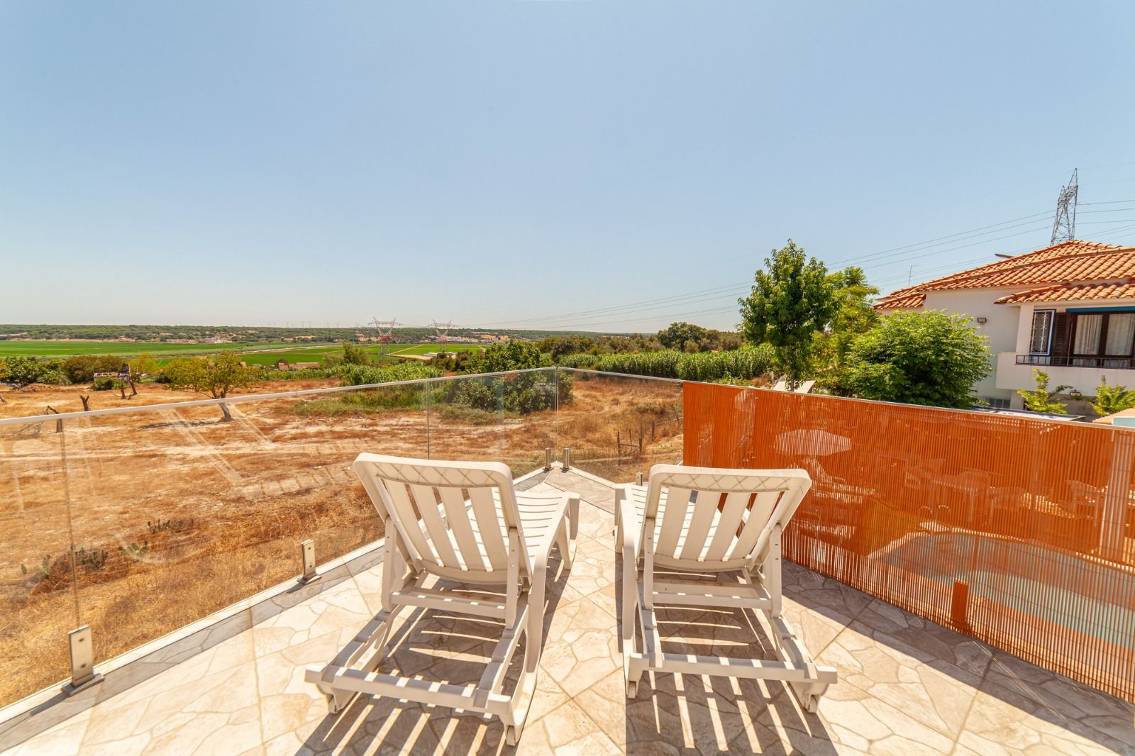 White patio chairs on a sunlit terrace overlooking open fields and a house with a red-tiled roof