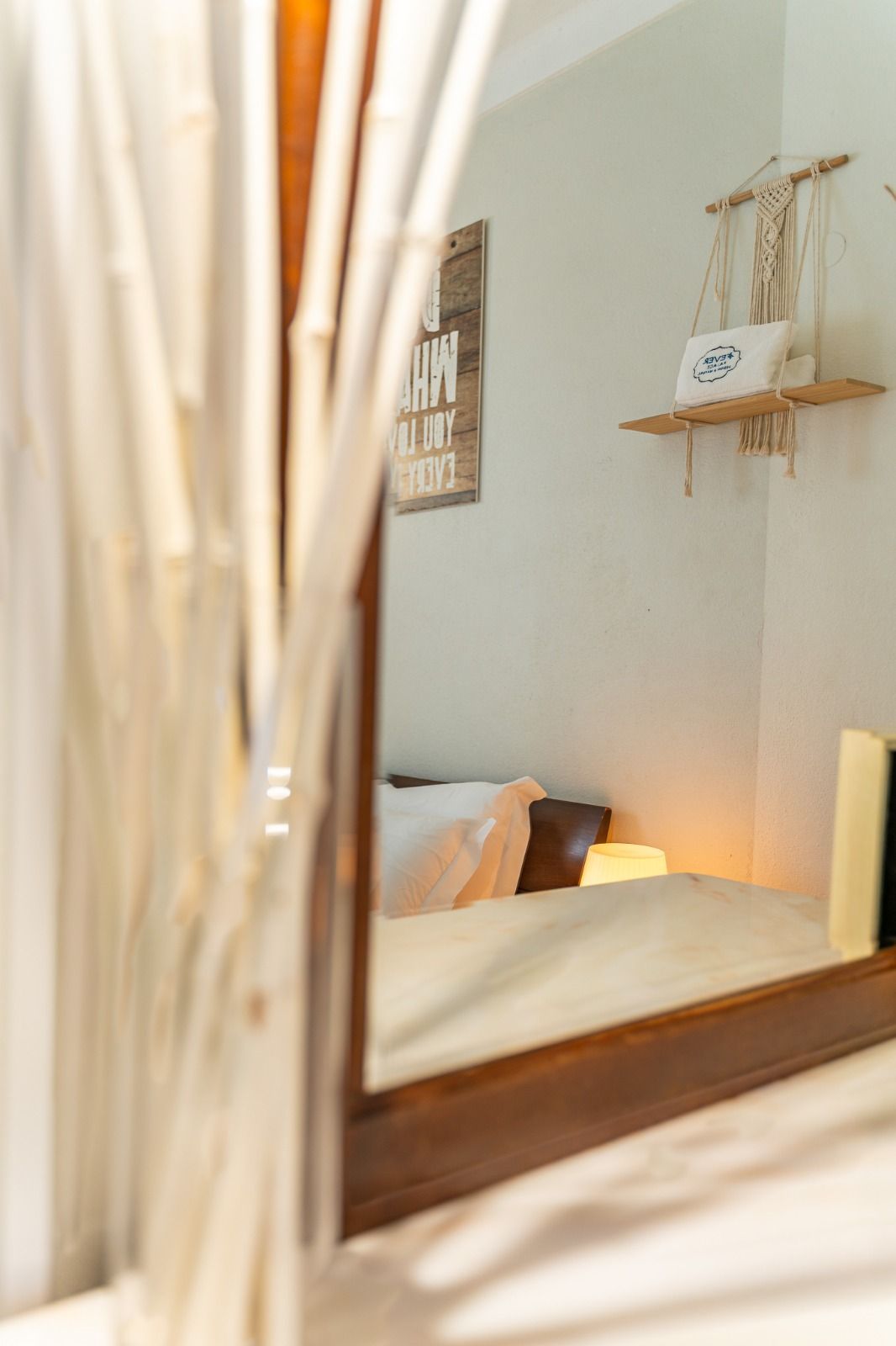 Cozy beige bedroom corner with a bed, warm lamp, wall shelf, and hanging decor, seen through curtains