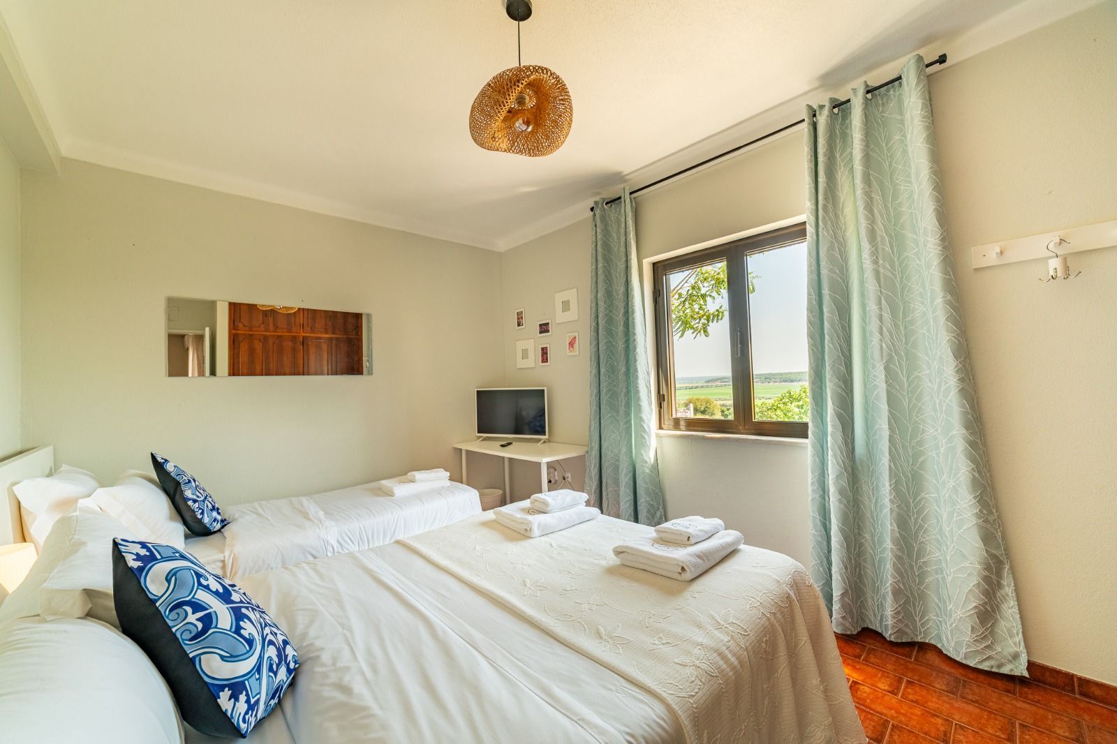 Bright bedroom with white bedding, blue pillows, wood floor, and large window with green curtains