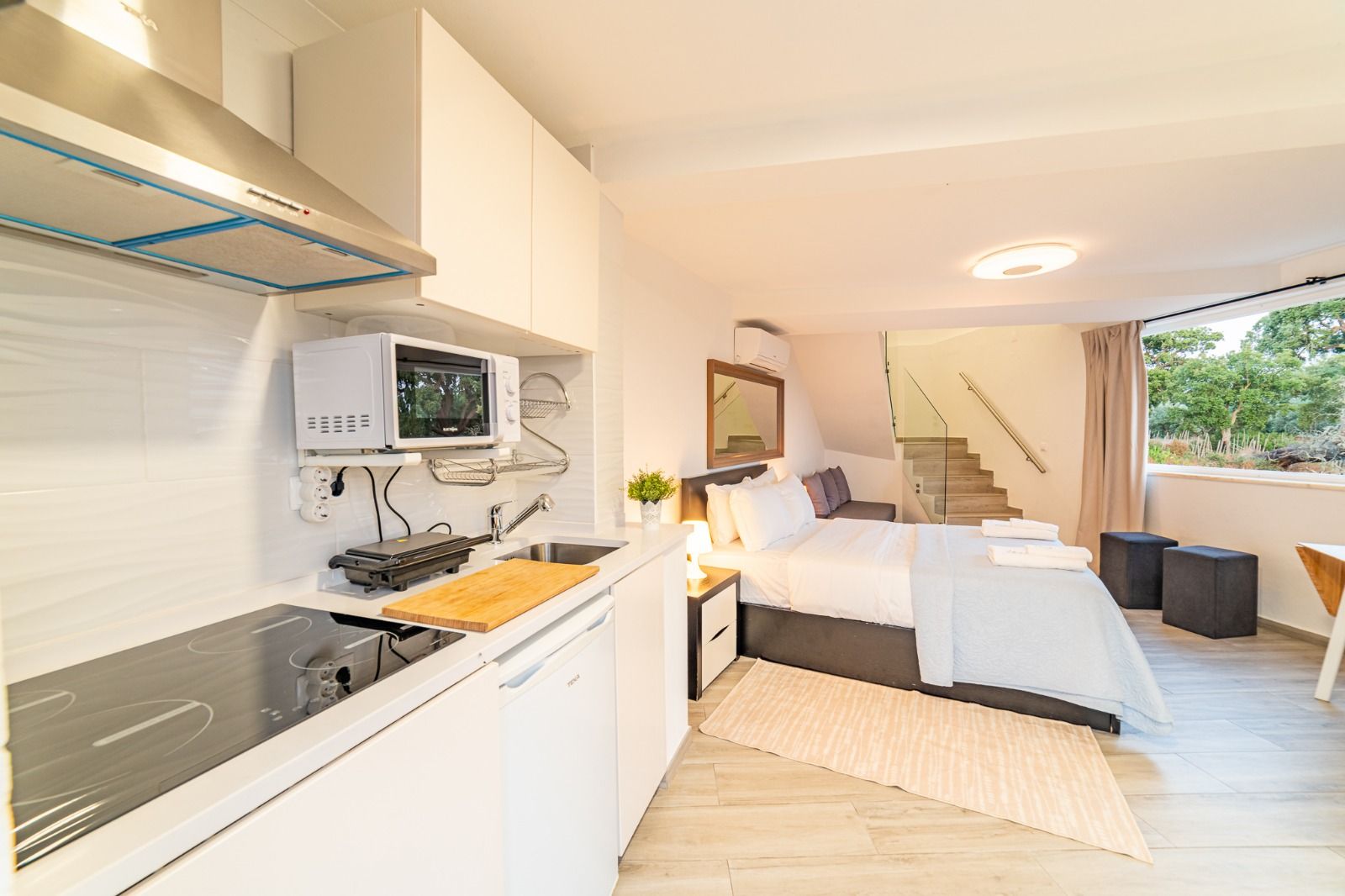 Bright studio with kitchenette, bed, and large windows overlooking greenery