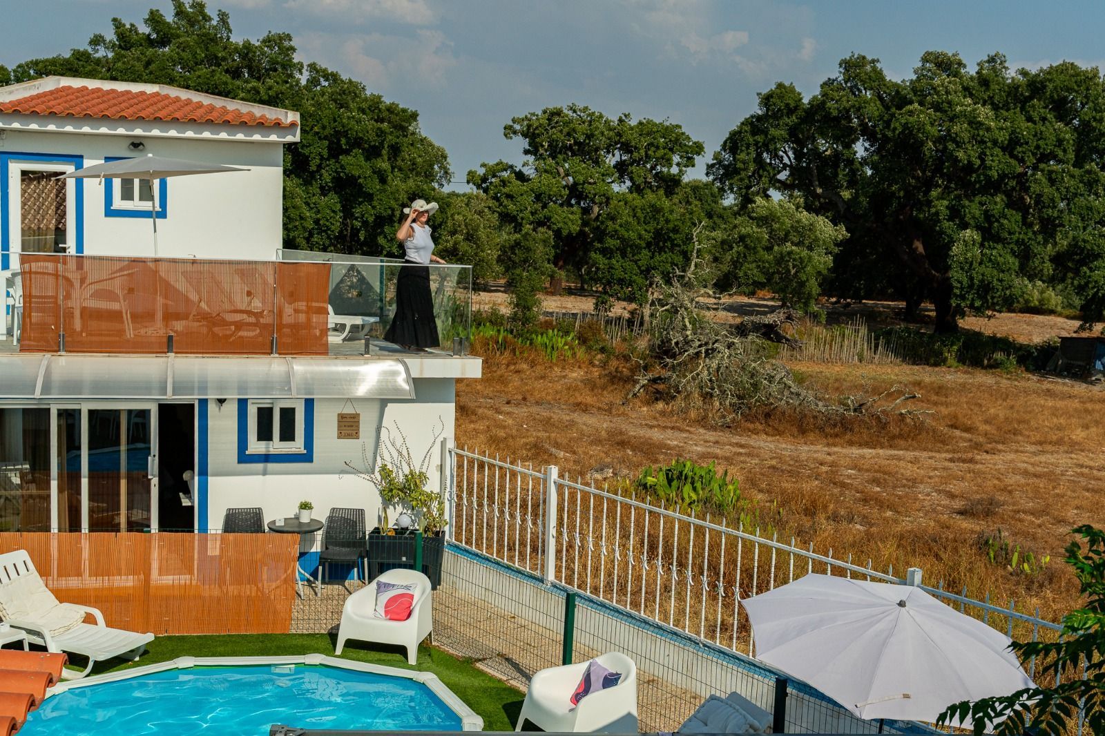 House with rooftop terrace and pool beside a dry field, with a person standing on the roof.