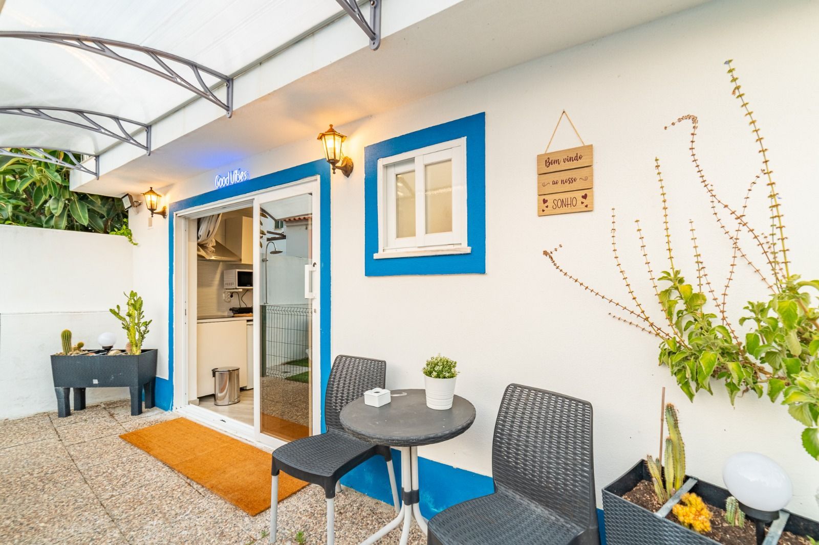 Small patio with blue-trimmed door, black chairs, round table, and potted plants under a white awning